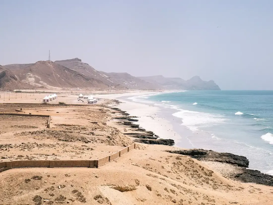 La lunga spiaggia di Al Mughsail, con onde bianche, mare azzurro e promontori rocciosi.
