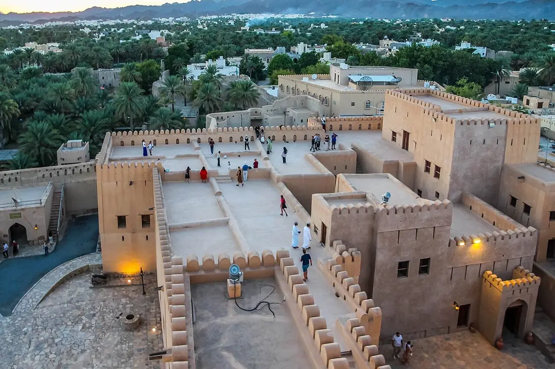 Cortili e mura merlate del forte di Nizwa, con il palmeto e le montagne sullo sfondo.