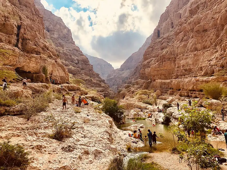 Pareti rocciose e una pozza naturale di Wadi Shab, con escursionisti lungo il sentiero.